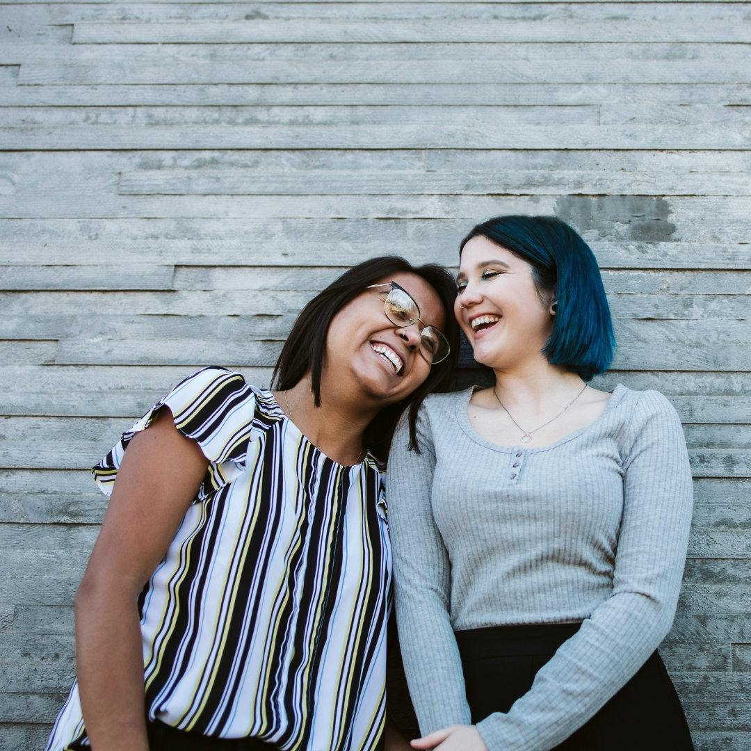 2 women smiling and standing beside gray wooden wall