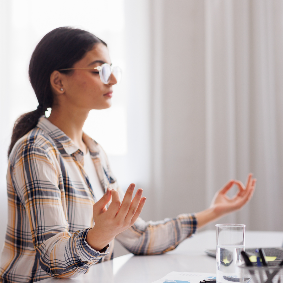 Managing stress in the workplace. Hindu student girl meditating and relaxing during a busy day while sitting at her workplace in an office interior. Focus on the hand.