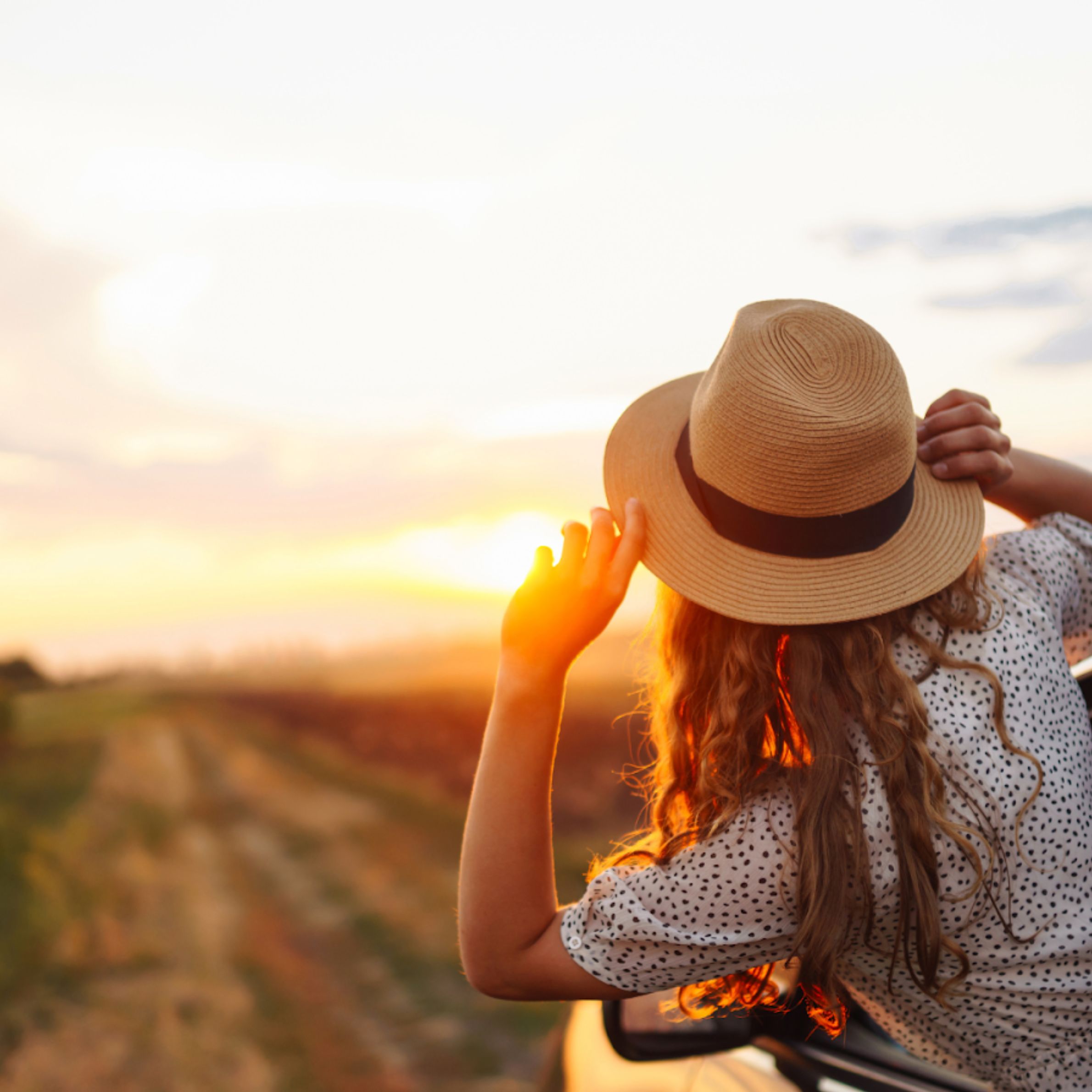 Woman wearing a hat looking at a sunset from a car window on a countryside road